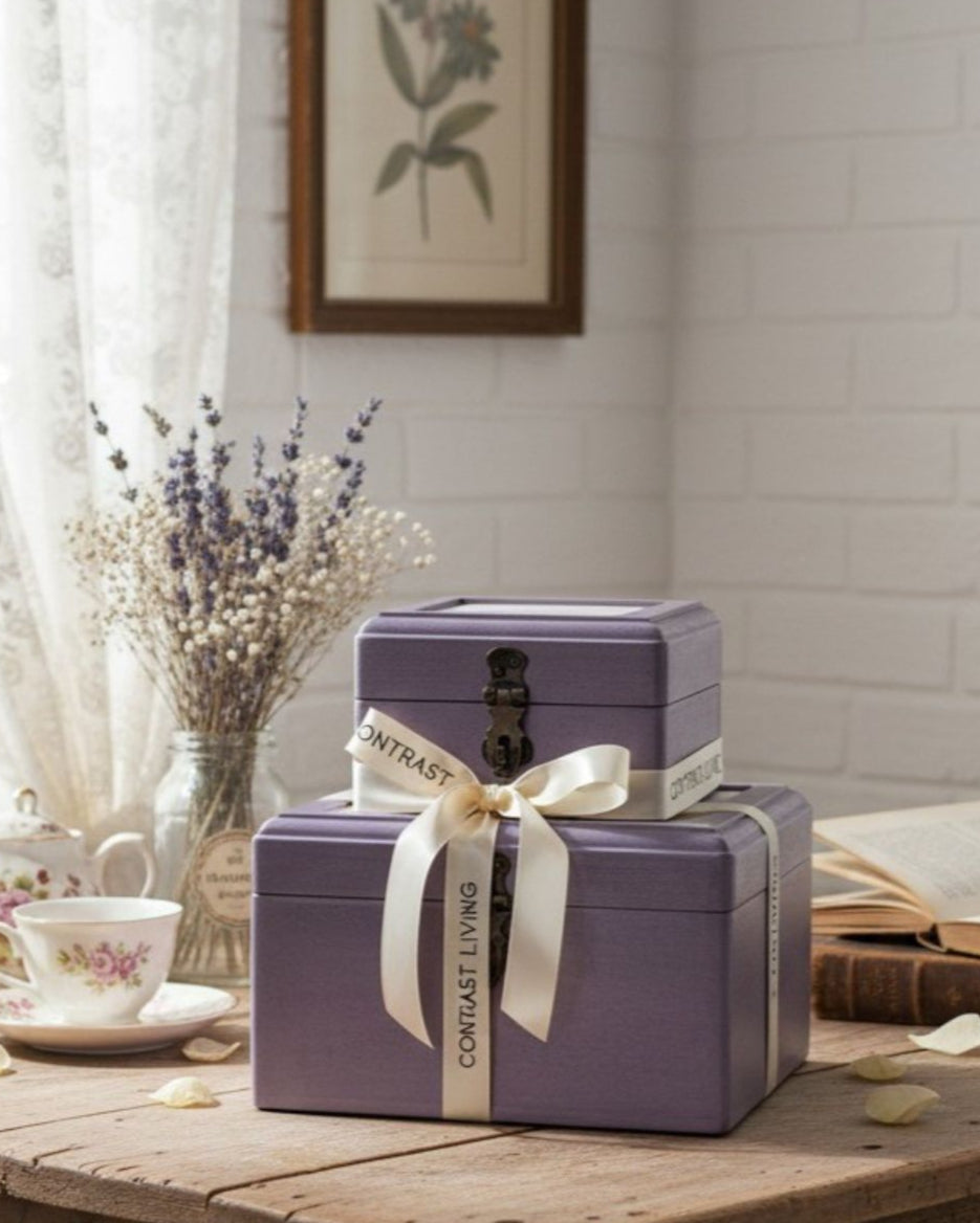 Stack of purple boxes with a white ribbon on a wooden table, with a floral painting in the background.